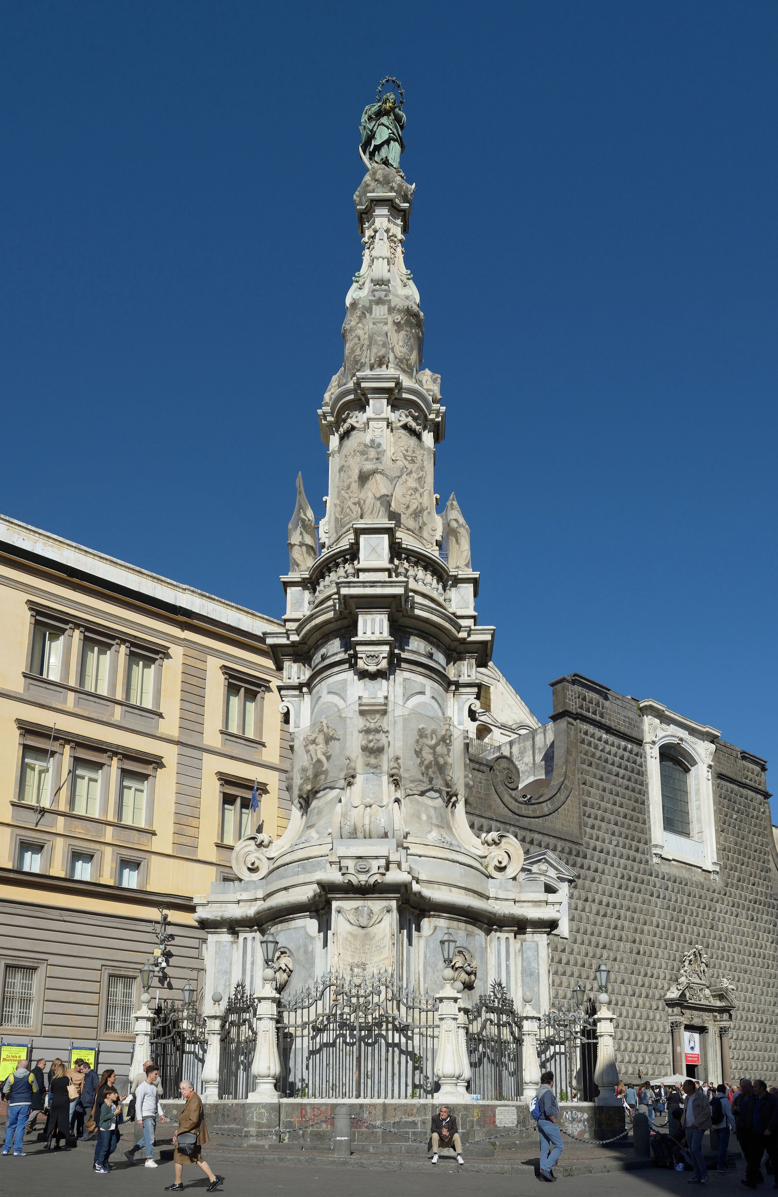 A historic monument with intricate carvings and statues, surrounded by classical architecture and a clear blue sky.