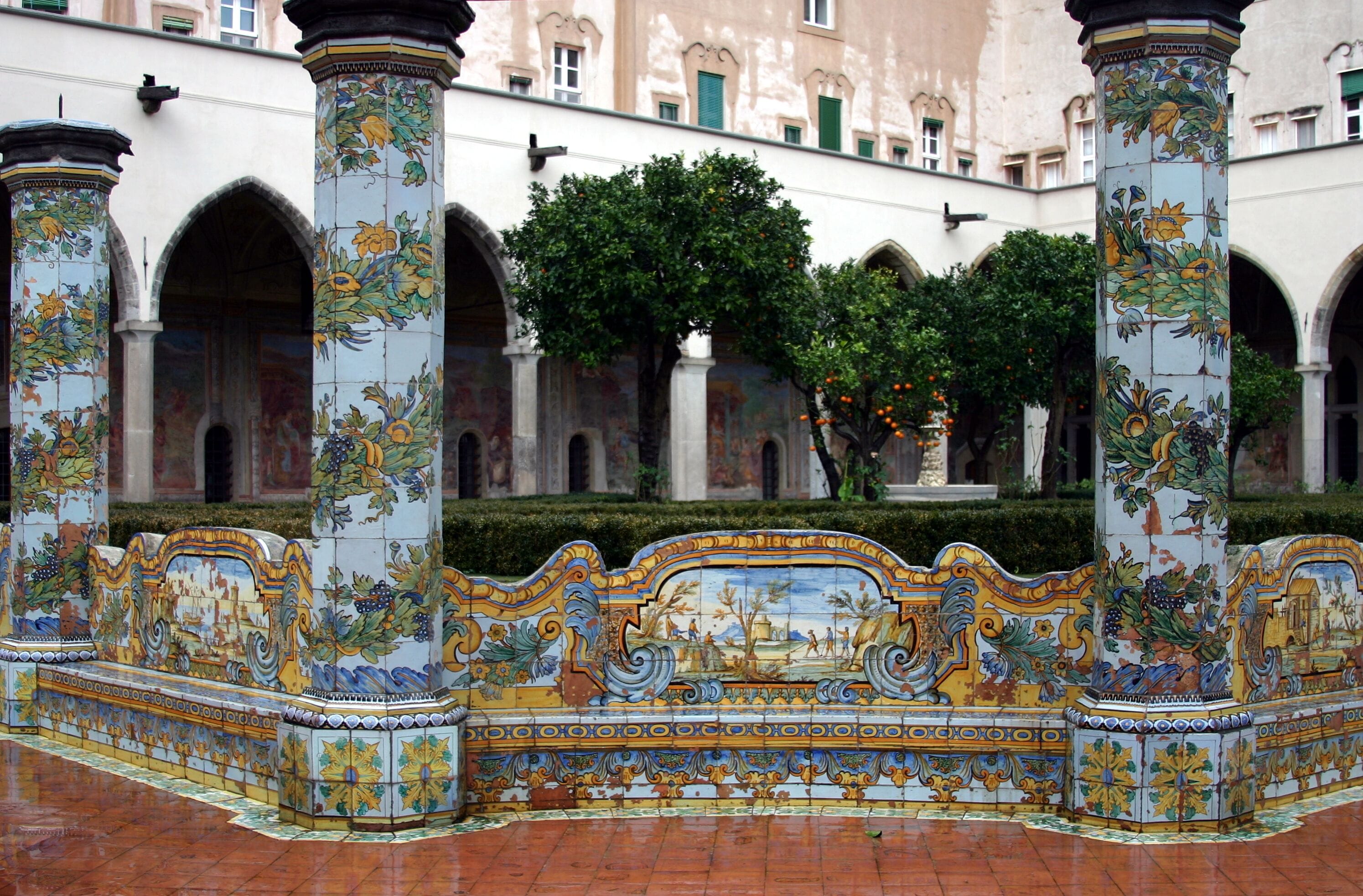 Un patio con columnas y arcos ricamente decorados con azulejos, rodeado de abundante vegetación.