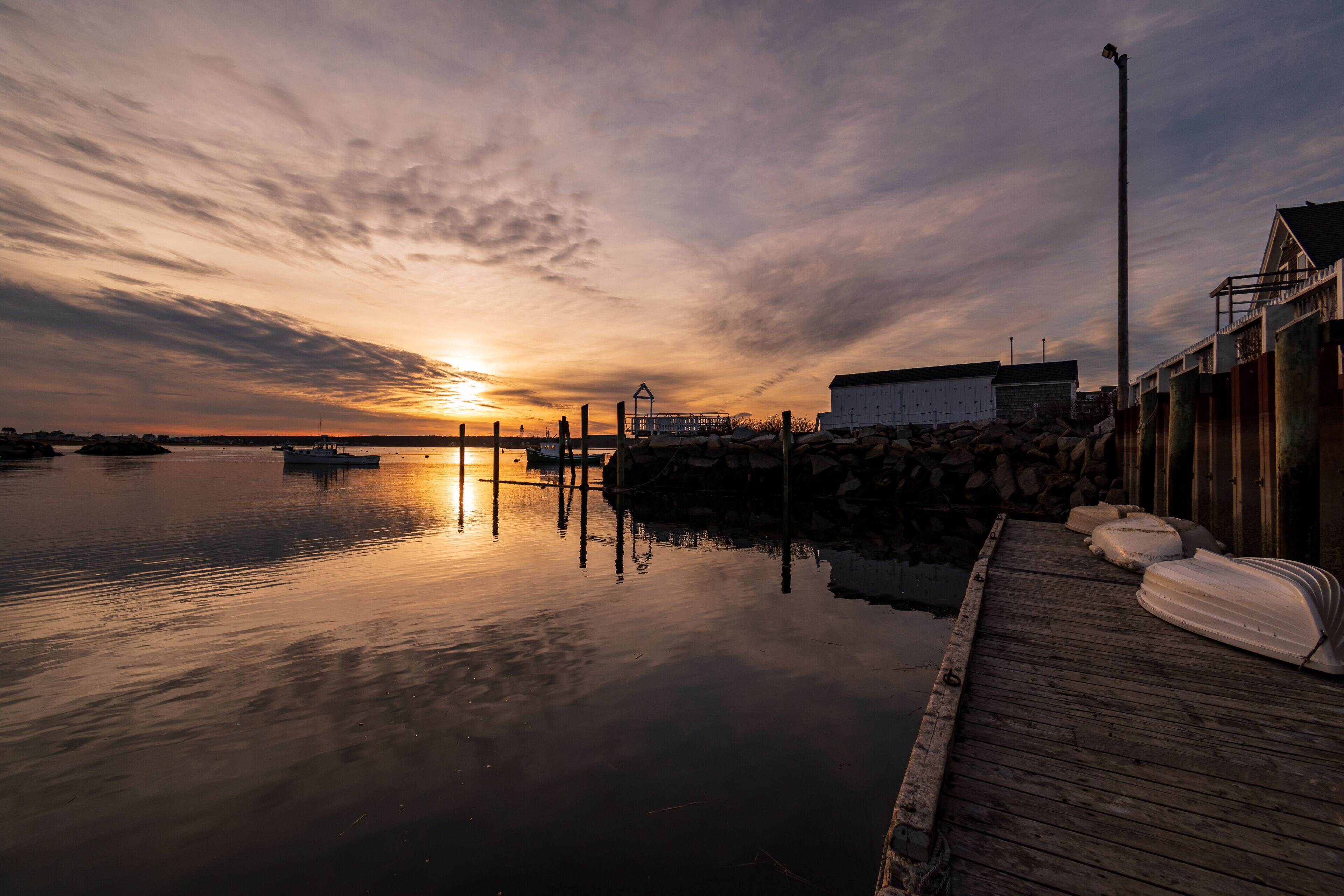 A sunset over a calm harbor with boats, a wooden dock, and a building in the background.