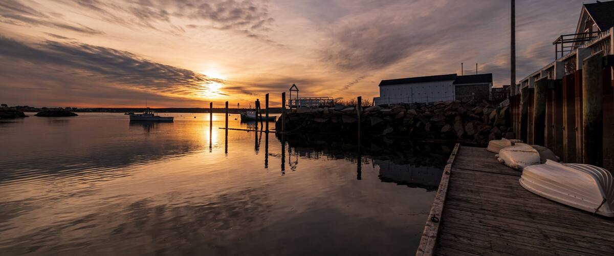 A sunset over a calm harbor with boats, a wooden dock, and a building in the background.