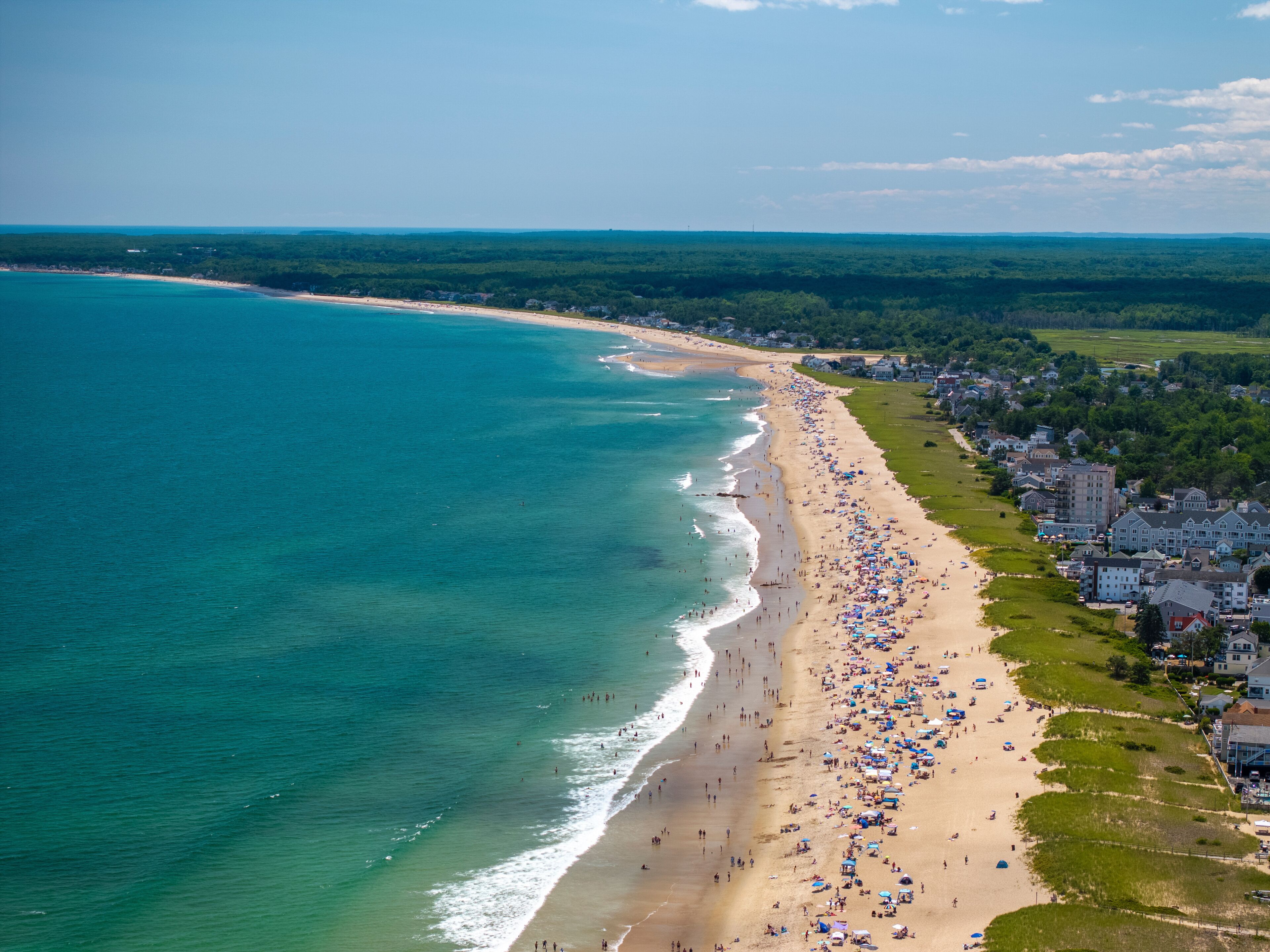 Eine Küstenstadt mit einem langen Sandstrand, blauem Meer und einem dichten Wald im Hintergrund.