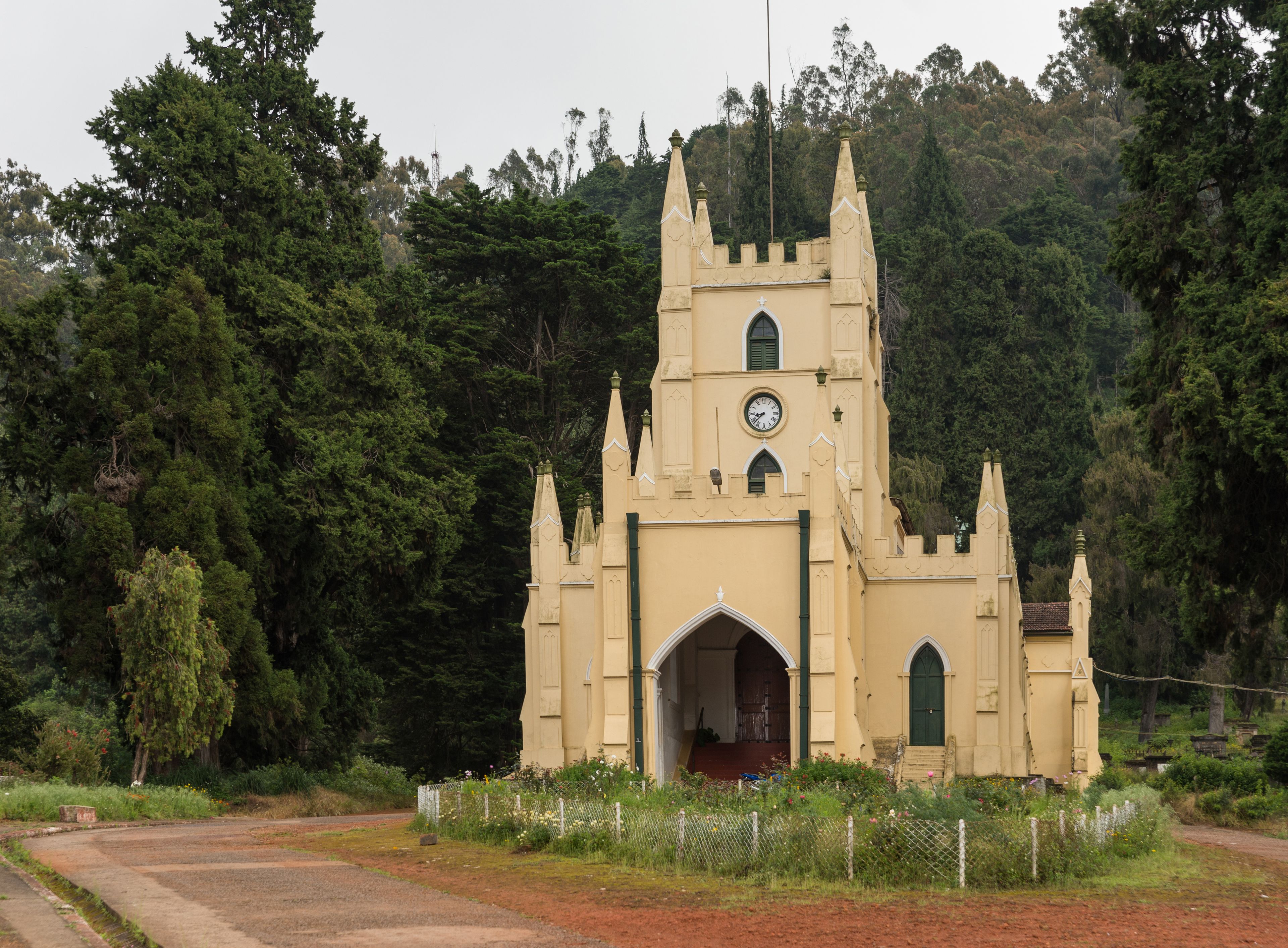 Ooty, India - October 25, 2013: The yellow painted castle-like Saint Stevens Church in Ooty. Red dirt and rose garden with dark green foliage in background. 