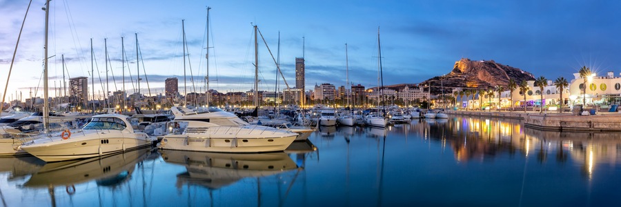 Una marina con varios yates amarrados, una colina destacada con una fortaleza y un paisaje urbano al fondo.