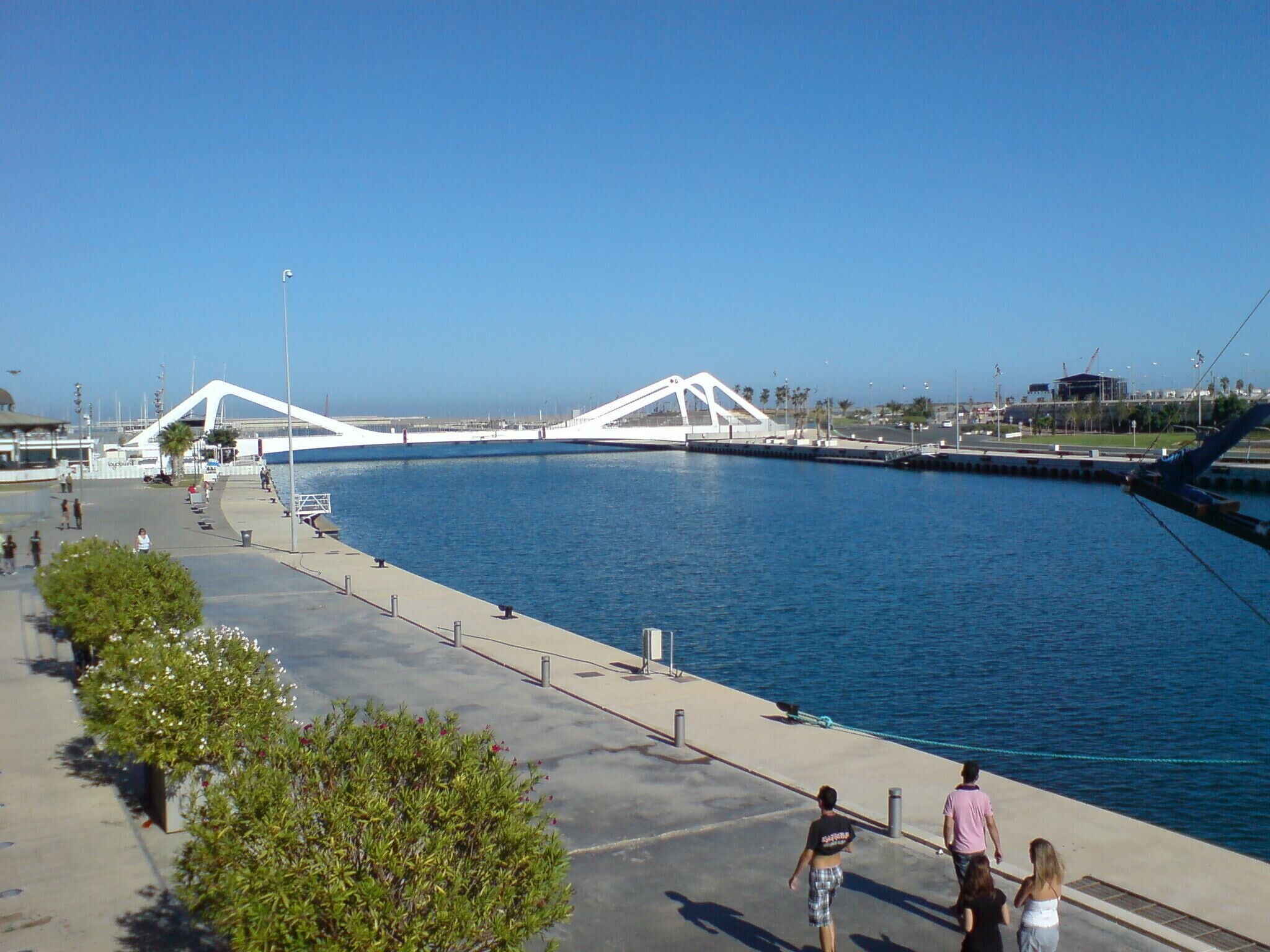 A waterfront promenade with a modern white bridge, a marina, and a clear blue sky.