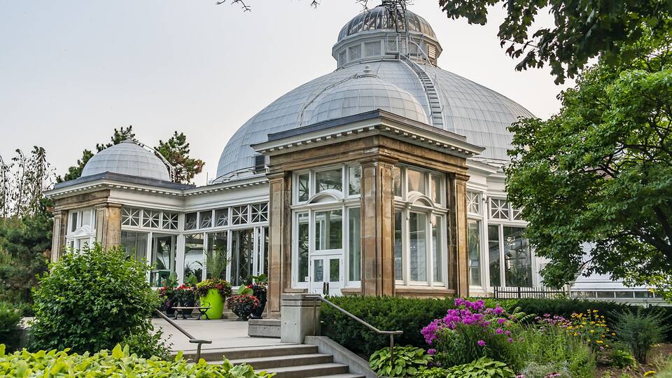 A large glass-domed building with a classical facade, surrounded by lush gardens and a clear sky.