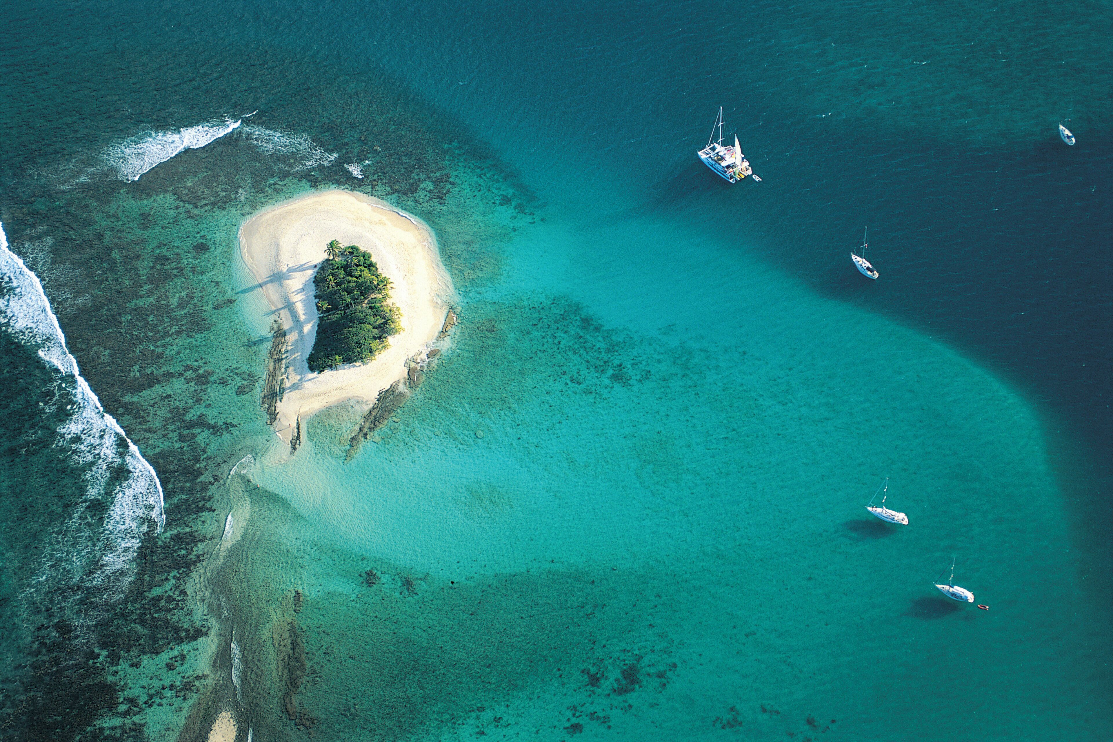 An aerial view of a turquoise lagoon with a small island, clear waters, and several boats.