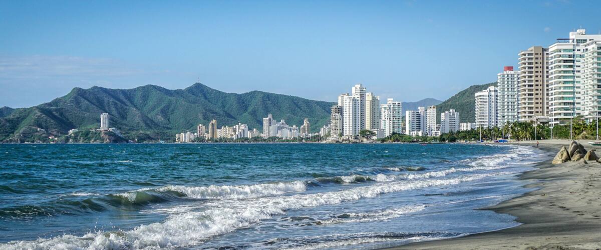 A beach with waves, a city skyline, and mountains in the background.