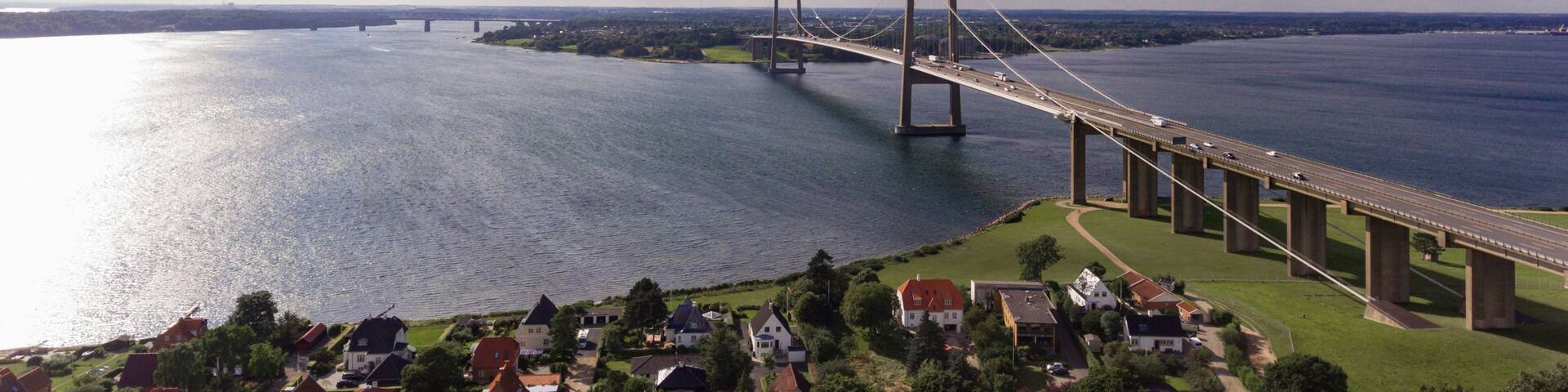 A large suspension bridge spanning a wide river, with a town and greenery in the foreground.