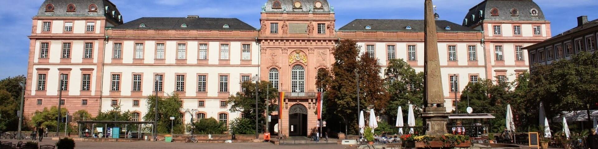 A historic building with a central entrance, a fountain, and surrounding trees.