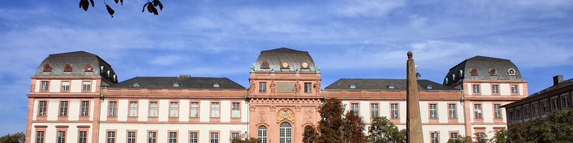 Un edificio histórico con entrada central, una fuente y árboles circundantes.