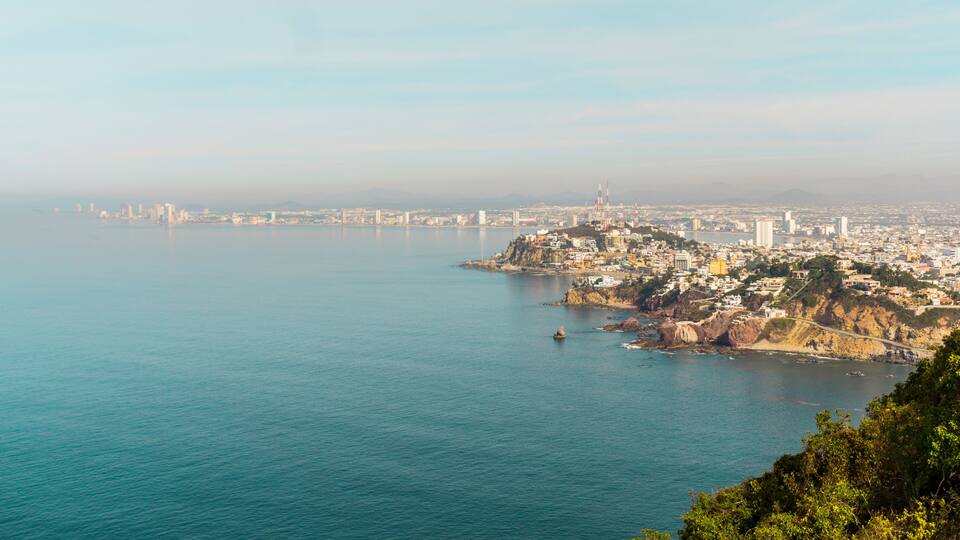 A coastal cityscape with a prominent bridge, a dense urban area, and a rocky coastline.