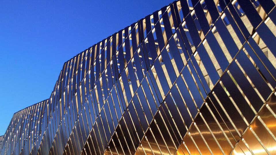Un bâtiment moderne doté d’une façade réfléchissante et d’un motif géométrique composé de lignes verticales et diagonales.