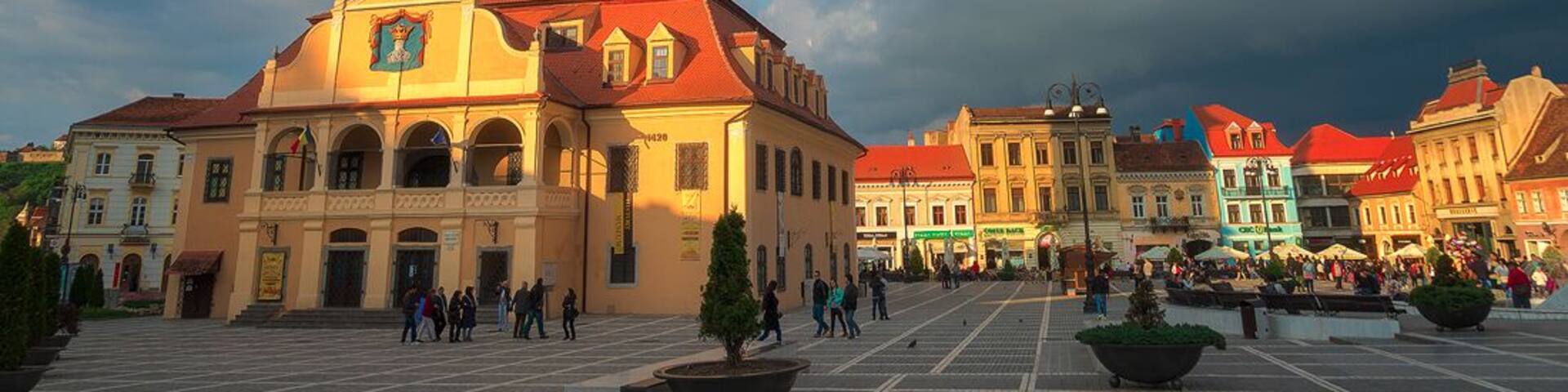 A historic building with a clock tower and red roof, surrounded by a square with people and trees.