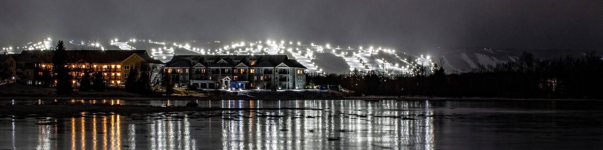 A nighttime view of a waterfront with illuminated buildings and a snowy landscape in the background.