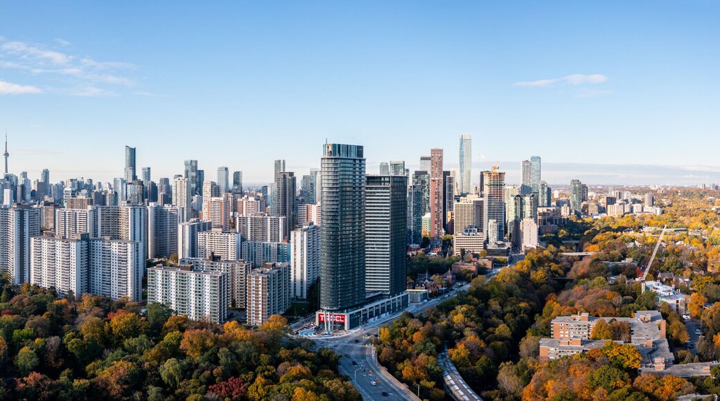 A stunning aerial view of Toronto's skyline in the fall, featuring vibrant autumn foliage, scattered clouds, and bright blue skies. The iconic cityscape is framed by golden and red-hued trees