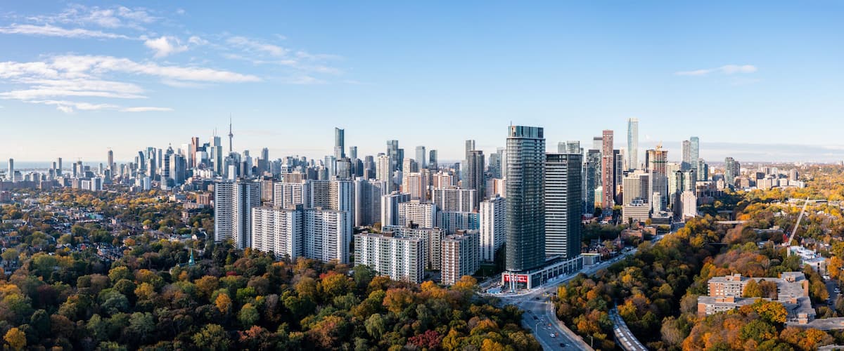 A stunning aerial view of Toronto's skyline in the fall, featuring vibrant autumn foliage, scattered clouds, and bright blue skies. The iconic cityscape is framed by golden and red-hued trees