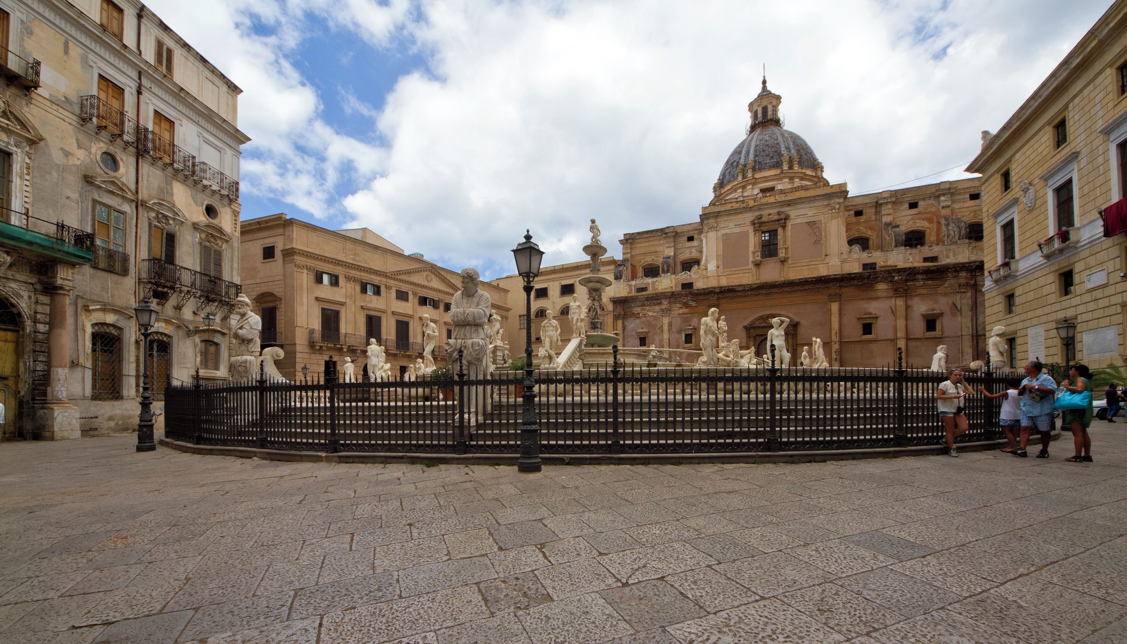 Una plaza histórica con una fuente, rodeada de arquitectura clásica y un edificio con cúpula.