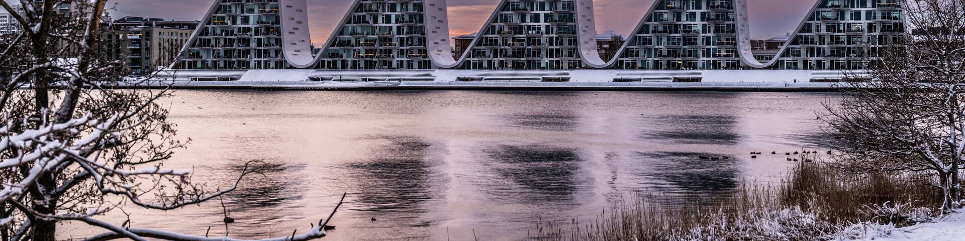 A modern architectural structure with a series of triangular peaks, a body of water in the foreground, and a snowy landscape.