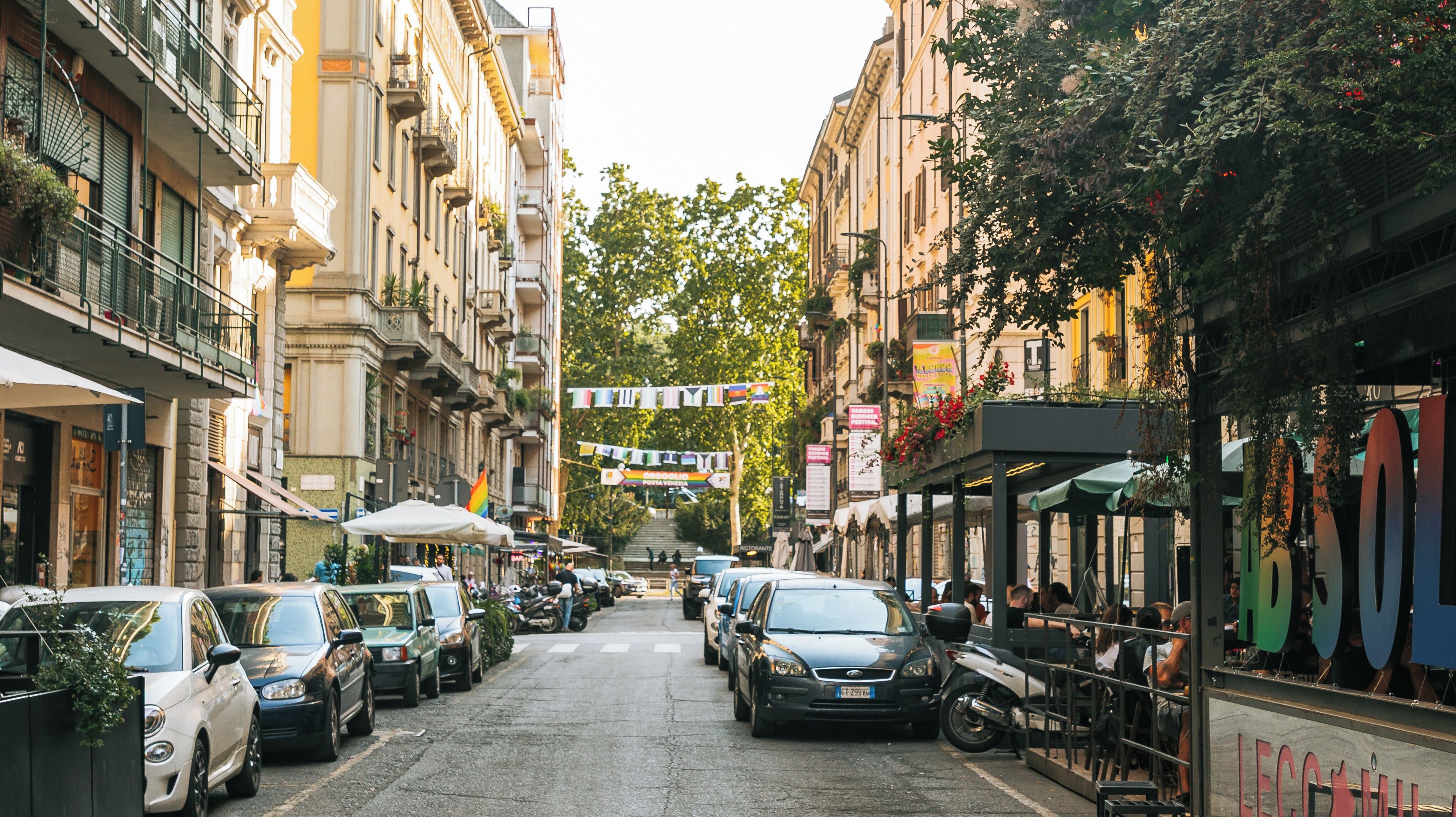 Une rue bordée de voitures garées et d’immeubles, avec un drapeau arc-en-ciel flottant entre les bâtiments.