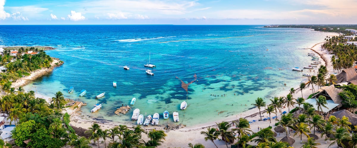 A coastal town with clear turquoise waters, boats, and a beach lined with palm trees and buildings.