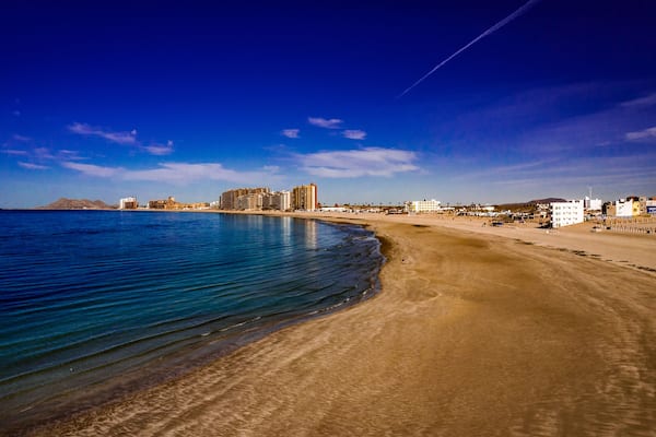 Un paisaje urbano costero con una playa de arena, mar tranquilo y cielo azul despejado.