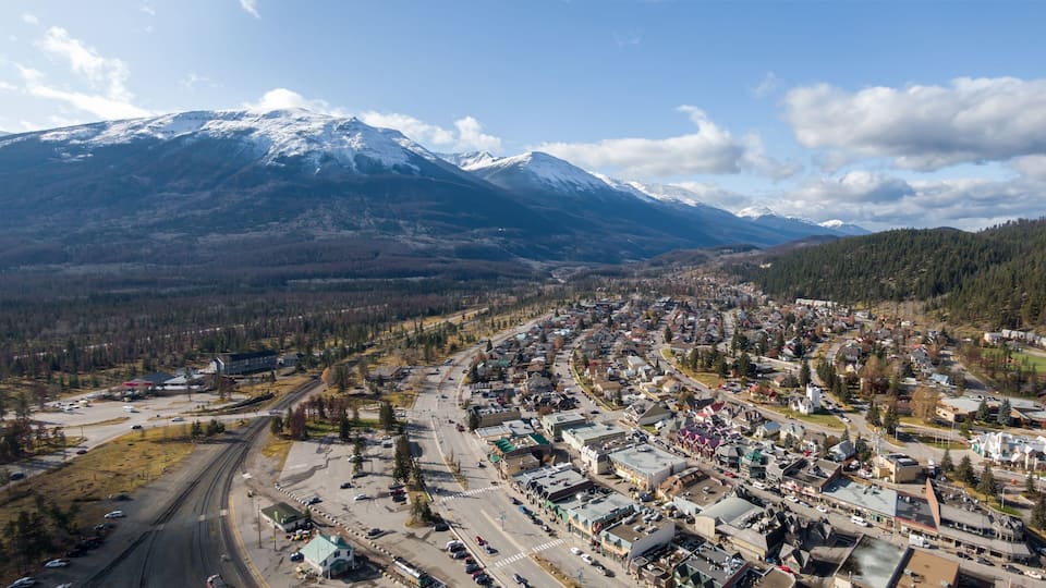 Aerial view of Jasper town nestled in the Canadian Rockies, with snow-capped peaks in the background. Trains and cars are visible in the town center. Alberta, Canada