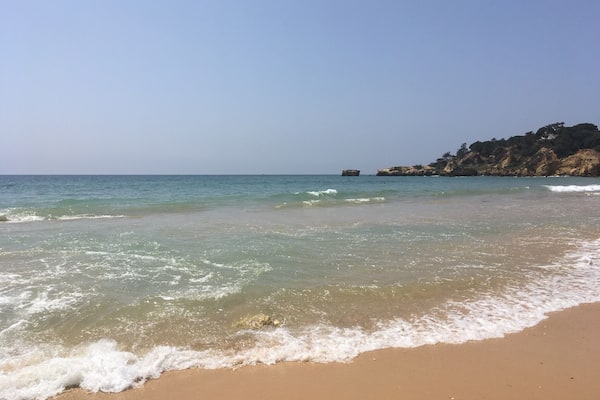 A beach with clear water, gentle waves, and rocky cliffs in the background.