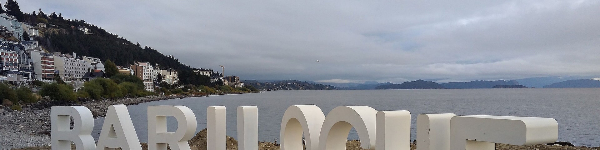 Large white letters spelling "BARILOCHE" on a wooden platform by a beach.