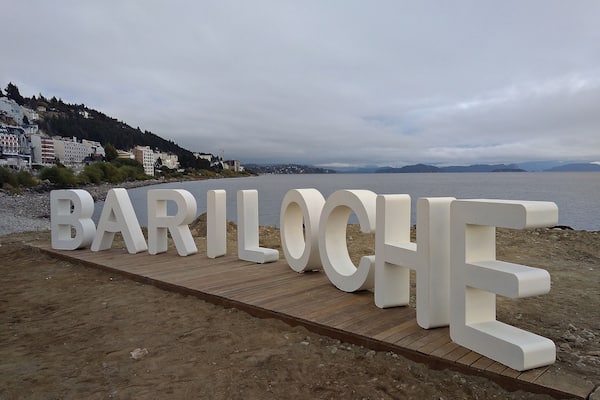 Large white letters spelling "BARILOCHE" on a wooden platform by a beach.