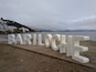 De grandes lettres blanches formant le mot « BARILOCHE » sont disposées sur une plateforme en bois, près d’une plage.