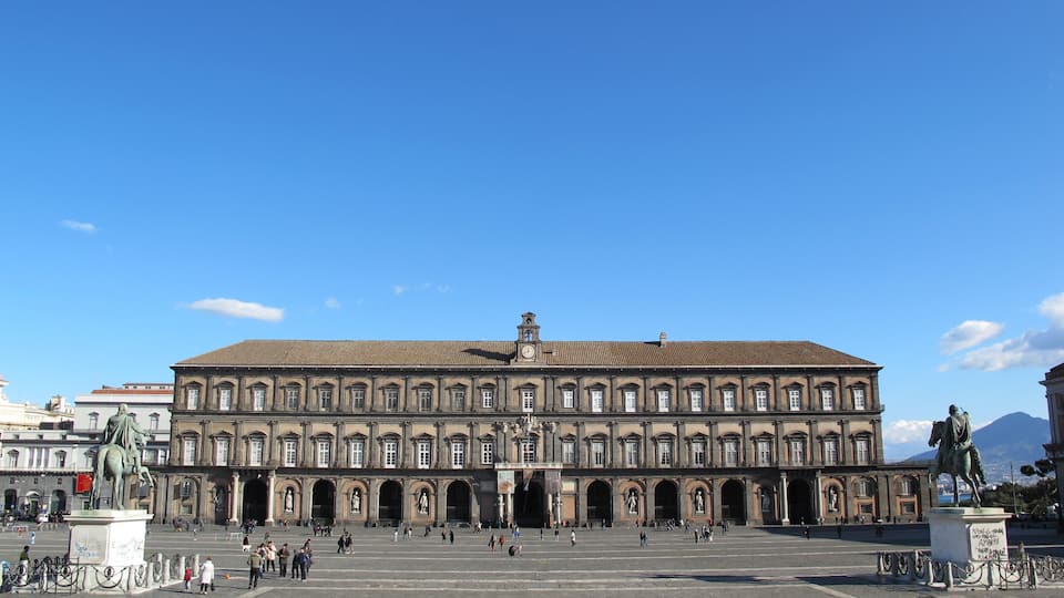 A large historic building with a central clock tower, surrounded by a plaza with statues and people.