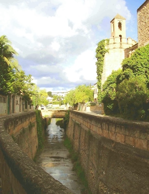 Un canal estrecho bordeado por muros de piedra, un puente a lo lejos y una torre con reloj.