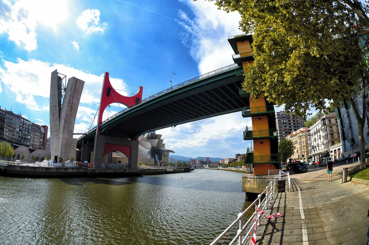 A modern bridge with a red sculpture, a river, and urban buildings.