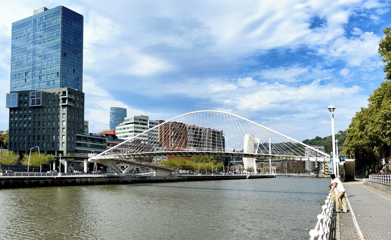 A modern bridge with a white arch over a river, surrounded by urban buildings and a clear sky.