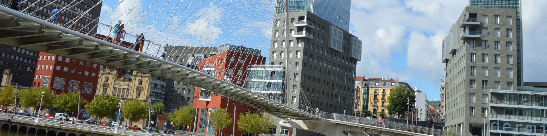 El Zubizuri (del euskera "puente blanco"), tambiĂ©n conocido como el Puente Peatonal del Campo de VolantĂn, aunque popularmente llamado puente de Calatrava, es un puente en arco (cuya pista cuelga de Ă©l) sobre la rĂa del NerviĂłn, en la ciudad vasca de Bilbao, en el norte de España. Une el Campo de VolantĂn (Castaños), en la margen derecha, con Uribitarte (el Ensanche), en la izquierda. Las torres que se ven detrĂĄs son las torres Isozaki que son parte de la "Puerta Isozaki" Isozaki Atea (en castellano, "Puerta Isozaki") es como se conoce a un complejo de siete edificios diseñado por el arquitecto japonĂ©s Arata Isozaki con la colaboraciĂłn del arquitecto bilbaĂno Iñaki Aurrekoetxea, en el distrito de Abando, en Bilbao, España. Se compone de dos torres gemelas de 82 metros y 23 plantas, y cinco edificios de entre 6 y 8 pisos. Tiene una superficie total de 84.000 mÂČ, de los cuales 8.200 mÂČ estĂĄn destinados a la actividad comercial, mientras que los restantes constituyen 317 viviendas, cuyos precios oscilan entre 400.000 y 800.000âŹ. En el complejo se asientan las sedes de Eusko Ikaskuntza, Unesco Etxea y el Instituto Europa de los Pueblos. DespuĂ©s de haberse intentado por diferentes iniciativas privadas rehabilitar el antiguo DepĂłsito Franco, situado en el Muelle de Uribitarte, y no habiĂ©ndose logrado este objetivo, se planteaba un importante y complejo problema urbanĂstico en Uribitarte. Por un lado, la pasarela Zubizuri diseñada por Santiago Calatrava no conecta exactamente con la zona del Ensanche, donde existe vida cotidiana intensa, creando una barrera fĂsica entre Ă©sta y la RĂa. Ăste es un problema genĂ©rico que existe en Bilbao, dado que la ciudad ha vivido siempre de espaldas a la RĂa. No existe conexiĂłn directa con la RĂa, motor de las actividades industriales de la ciudad. Por otro lado, el Ayuntamiento lleva ya varios años con el intento de crear elementos importantes a lo largo de la RĂa. Claros exponentes de este criterio son puntos como el Museo Guggenheim Bilbao, el Paseo de Abandoibarra o el Palacio Euskalduna y las demĂĄs obras de la Sociedad Bilbao RĂa 2000, con el fin de dirigir la recuperaciĂłn y transformaciĂłn de las zonas degradadas de Bilbao y su ĂĄrea metropolitana. AsĂ, sumĂĄndose a estas construcciones, se pretendĂa tambiĂ©n la construcciĂłn de una plaza pĂșblica sobre la colindante calle Marina, y resolver asĂ la problemĂĄtica de un desnivel de unos 14 metros entre la RĂa y la zona del Ensanche. Posteriormente, la empresa promotora Vizcaina de Edificaciones de Grupo Urvasco S.A. gestionĂł con los propietarios del solar sito al otro lado de la calle Marina la realizaciĂłn de una actuaciĂłn conjunta del ĂĄrea, y el proyecto de la plaza quedĂł atrĂĄs. La nueva soluciĂłn, expuesta por el arquitecto japonĂ©s Arata Isozaki fue diseñar una Gran Escalinata que tiene una anchura de unos 50 metros, en el encuentro con el Paseo de Uribitarte, en equivalencia a la de la Plaza de España en Roma, para conectar estos dos niveles y hacer dos edificios de 23 plantas, unos 82 metros de altura, a ambos lados de la escalinata. Era la propuesta de diseñar una Puerta a Bilbao en el siglo XXI con el fin de conectar el Ensanche y la RĂa. De ahĂ el nombre de las torres, Isozaki Atea, que en euskera significa Puerta Isozaki. Con las torres ya construidas, la elecciĂłn de diferentes acabados pretende formar un conjunto diversificado y diferenciador respecto a las viviendas masificadas habituales. Se ha empleado vidrio en la fachada de las torres y aplacado de piedras y ladrillo visto o vidrios en los bloques restantes, diferenciando los diferentes volĂșmenes que componen el conjunto con la textura del material.