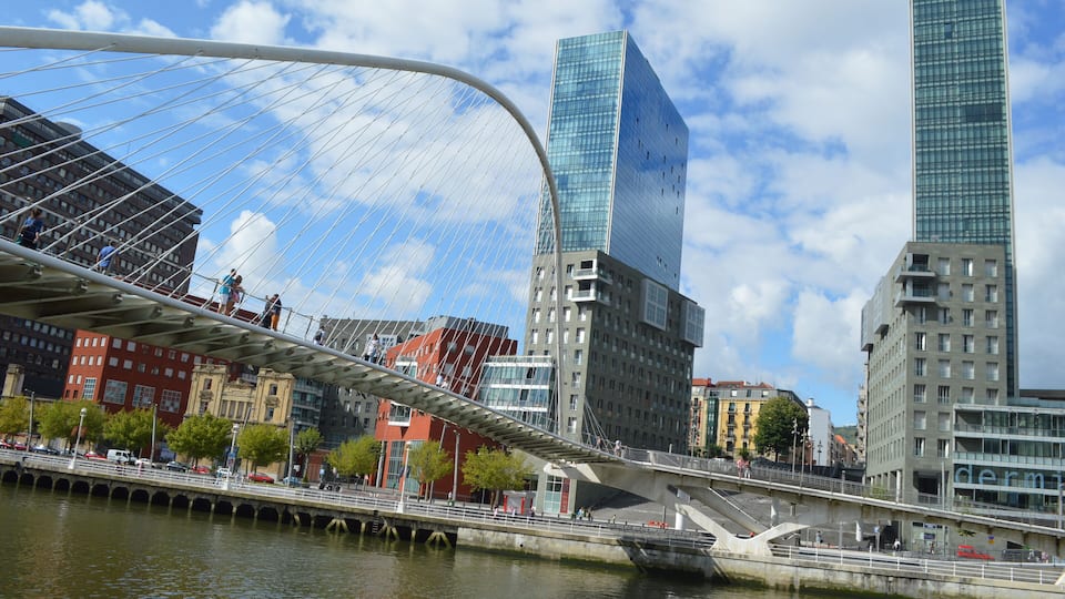 El Zubizuri (del euskera "puente blanco"), también conocido como el Puente Peatonal del Campo de Volantín, aunque popularmente llamado puente de Calatrava, es un puente en arco (cuya pista cuelga de él) sobre la ría del Nervión, en la ciudad vasca de Bilbao, en el norte de España. Une el Campo de Volantín (Castaños), en la margen derecha, con Uribitarte (el Ensanche), en la izquierda. Las torres que se ven detrás son las torres Isozaki que son parte de la "Puerta Isozaki" Isozaki Atea (en castellano, "Puerta Isozaki") es como se conoce a un complejo de siete edificios diseñado por el arquitecto japonés Arata Isozaki con la colaboración del arquitecto bilbaíno Iñaki Aurrekoetxea, en el distrito de Abando, en Bilbao, España. Se compone de dos torres gemelas de 82 metros y 23 plantas, y cinco edificios de entre 6 y 8 pisos. Tiene una superficie total de 84.000 m², de los cuales 8.200 m² están destinados a la actividad comercial, mientras que los restantes constituyen 317 viviendas, cuyos precios oscilan entre 400.000 y 800.000€. En el complejo se asientan las sedes de Eusko Ikaskuntza, Unesco Etxea y el Instituto Europa de los Pueblos. Después de haberse intentado por diferentes iniciativas privadas rehabilitar el antiguo Depósito Franco, situado en el Muelle de Uribitarte, y no habiéndose logrado este objetivo, se planteaba un importante y complejo problema urbanístico en Uribitarte. Por un lado, la pasarela Zubizuri diseñada por Santiago Calatrava no conecta exactamente con la zona del Ensanche, donde existe vida cotidiana intensa, creando una barrera física entre ésta y la Ría. Éste es un problema genérico que existe en Bilbao, dado que la ciudad ha vivido siempre de espaldas a la Ría. No existe conexión directa con la Ría, motor de las actividades industriales de la ciudad. Por otro lado, el Ayuntamiento lleva ya varios años con el intento de crear elementos importantes a lo largo de la Ría. Claros exponentes de este criterio son puntos como el Museo Guggenheim Bilbao, el Paseo de Abandoibarra o el Palacio Euskalduna y las demás obras de la Sociedad Bilbao Ría 2000, con el fin de dirigir la recuperación y transformación de las zonas degradadas de Bilbao y su área metropolitana. Así, sumándose a estas construcciones, se pretendía también la construcción de una plaza pública sobre la colindante calle Marina, y resolver así la problemática de un desnivel de unos 14 metros entre la Ría y la zona del Ensanche. Posteriormente, la empresa promotora Vizcaina de Edificaciones de Grupo Urvasco S.A. gestionó con los propietarios del solar sito al otro lado de la calle Marina la realización de una actuación conjunta del área, y el proyecto de la plaza quedó atrás. La nueva solución, expuesta por el arquitecto japonés Arata Isozaki fue diseñar una Gran Escalinata que tiene una anchura de unos 50 metros, en el encuentro con el Paseo de Uribitarte, en equivalencia a la de la Plaza de España en Roma, para conectar estos dos niveles y hacer dos edificios de 23 plantas, unos 82 metros de altura, a ambos lados de la escalinata. Era la propuesta de diseñar una Puerta a Bilbao en el siglo XXI con el fin de conectar el Ensanche y la Ría. De ahí el nombre de las torres, Isozaki Atea, que en euskera significa Puerta Isozaki. Con las torres ya construidas, la elección de diferentes acabados pretende formar un conjunto diversificado y diferenciador respecto a las viviendas masificadas habituales. Se ha empleado vidrio en la fachada de las torres y aplacado de piedras y ladrillo visto o vidrios en los bloques restantes, diferenciando los diferentes volúmenes que componen el conjunto con la textura del material.