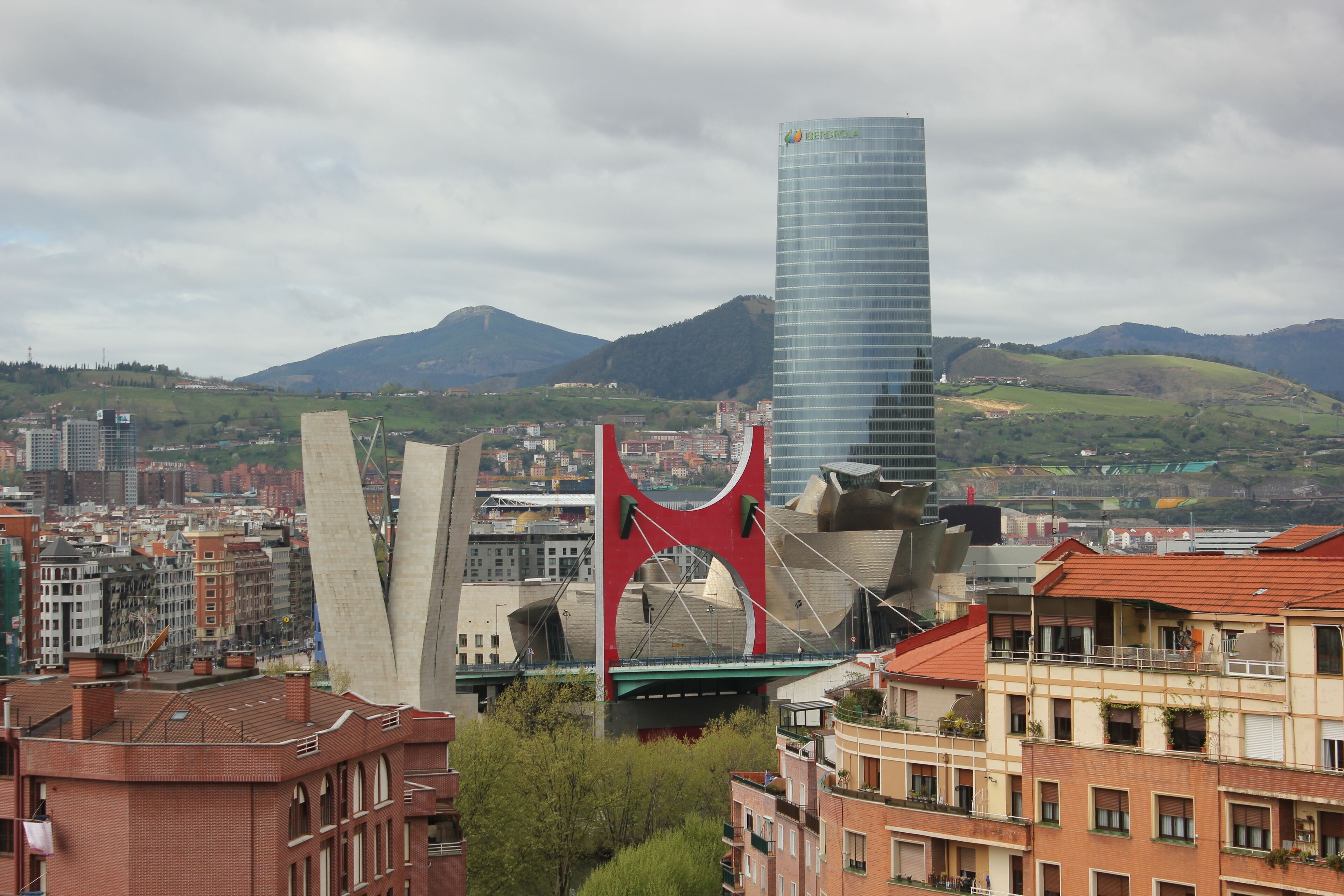 A cityscape with a prominent red bridge, a tall modern building, and a mountainous backdrop.