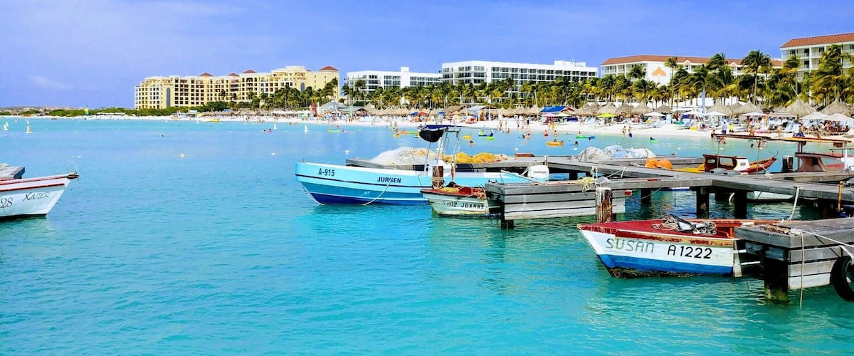 A marina with several boats docked, a beach with people, and a resort in the background.
