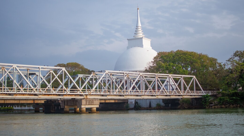 A white pagoda with a pointed spire, a bridge with a metal truss structure, and a body of water with trees in the background.
