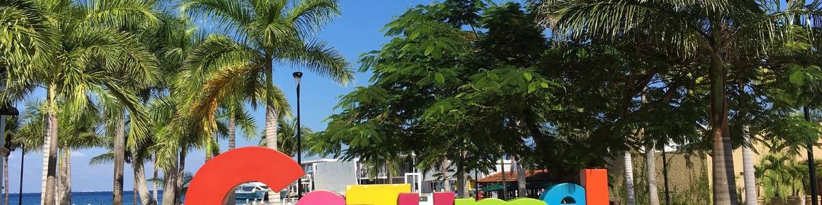 Une enseigne colorée sur laquelle on peut lire « ISLA COZUMEL », entourée de palmiers et surplombée par un ciel bleu clair.