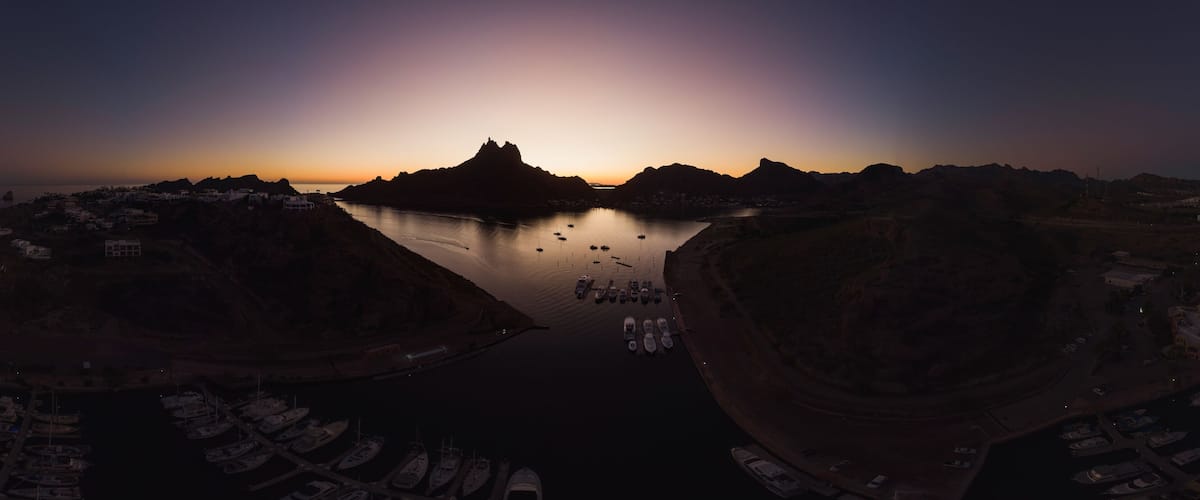 A coastal landscape with a marina, boats, and a prominent mountain at sunset.