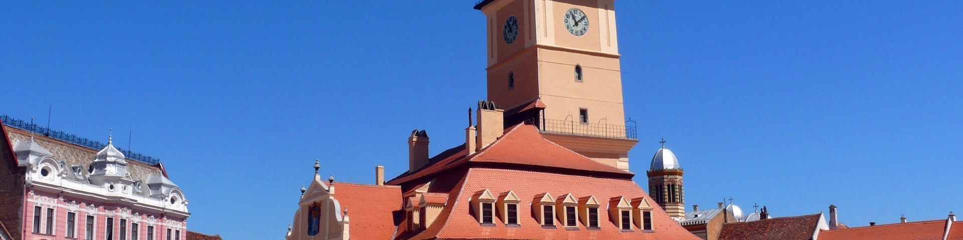 Um edifício histórico com uma torre de relógio, cercado por uma praça com bancos e uma fonte.