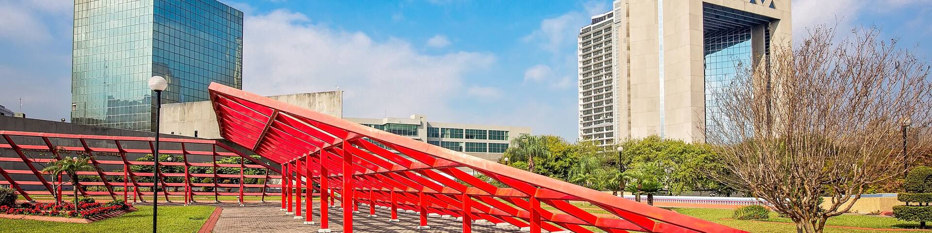 A modern urban park with a red metal sculpture, a pathway, and tall buildings in the background.