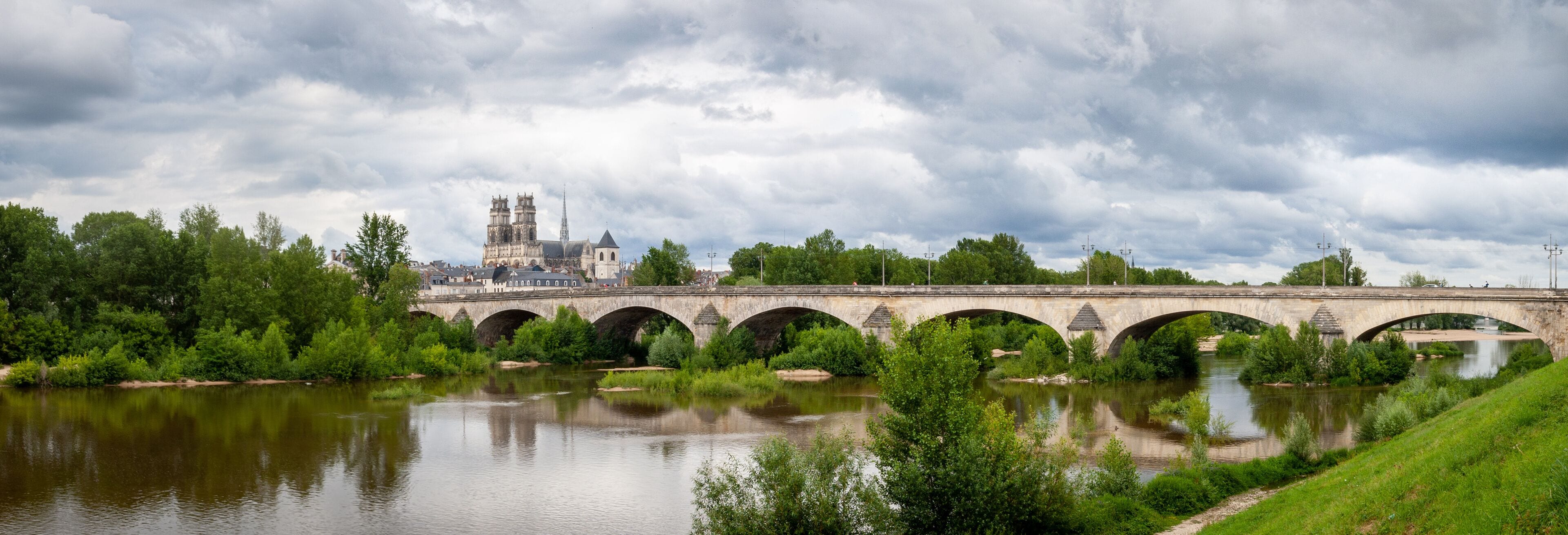 A stone bridge over a river with lush greenery and a historic building in the background.