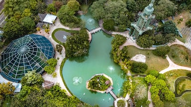 An aerial view of a park with a large green pond, a glasshouse, and a tower.