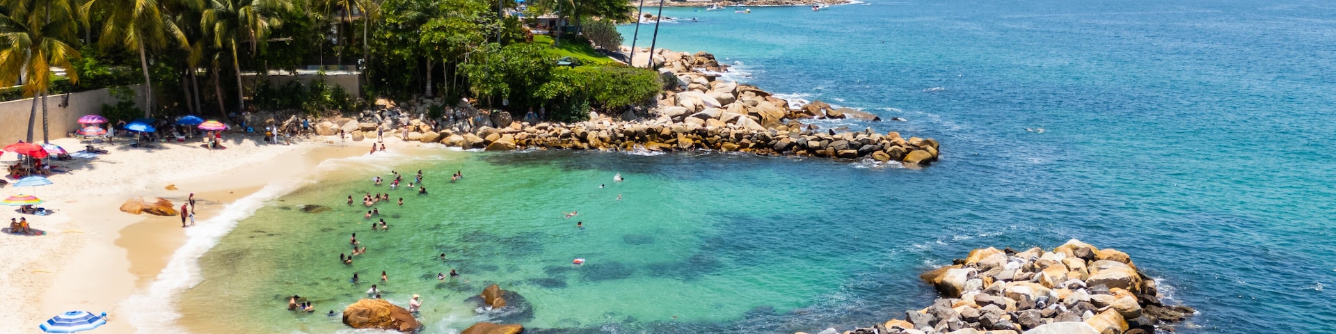 A tropical beach with clear turquoise waters, rocky shorelines, palm trees, and a hotel in the background.