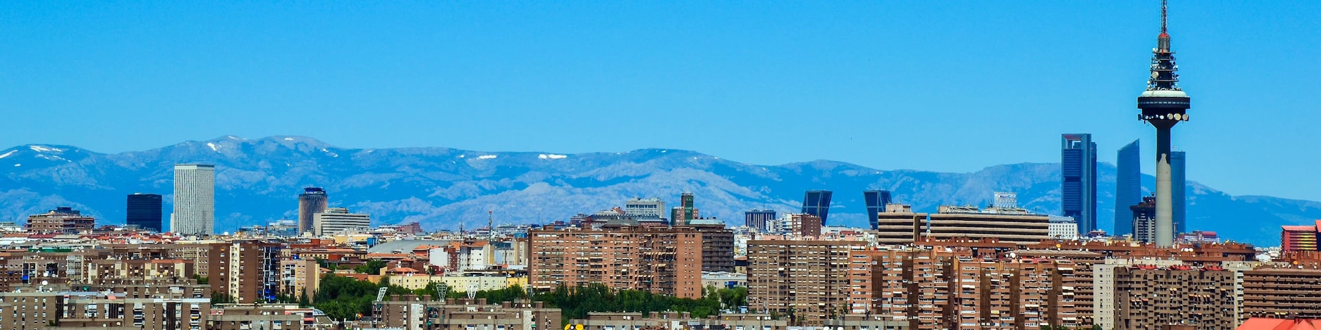 Paisagem urbana com edifícios altos e uma torre imponente ao fundo, com montanhas.
