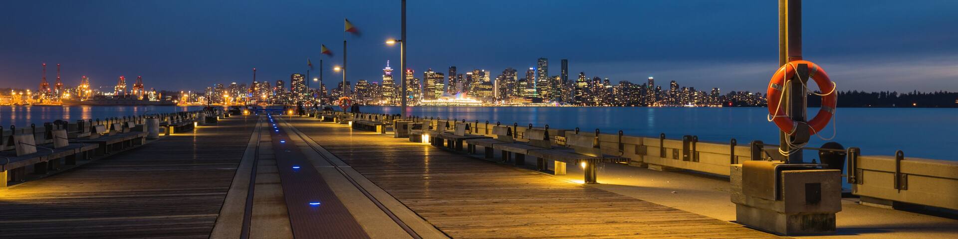 A waterfront promenade at dusk with city lights in the background.