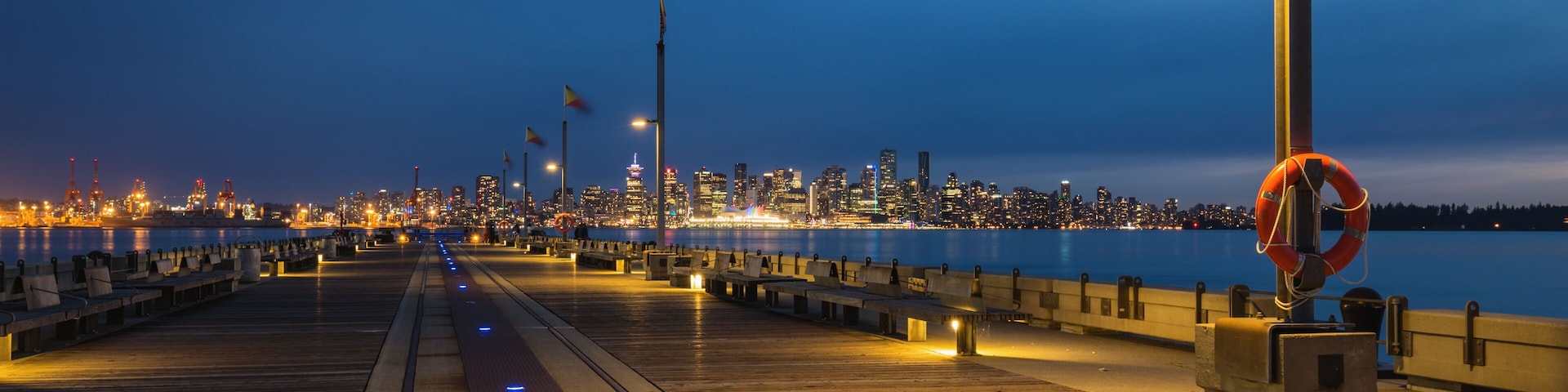 A waterfront promenade at dusk with city lights in the background.