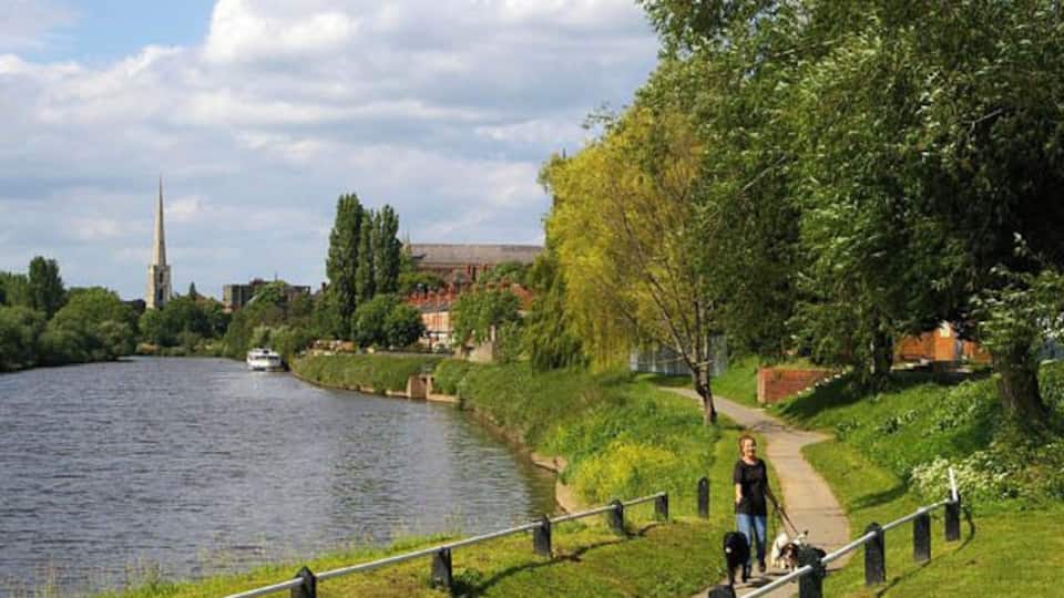 A river with a boat, a pedestrian path, a church spire, and greenery.